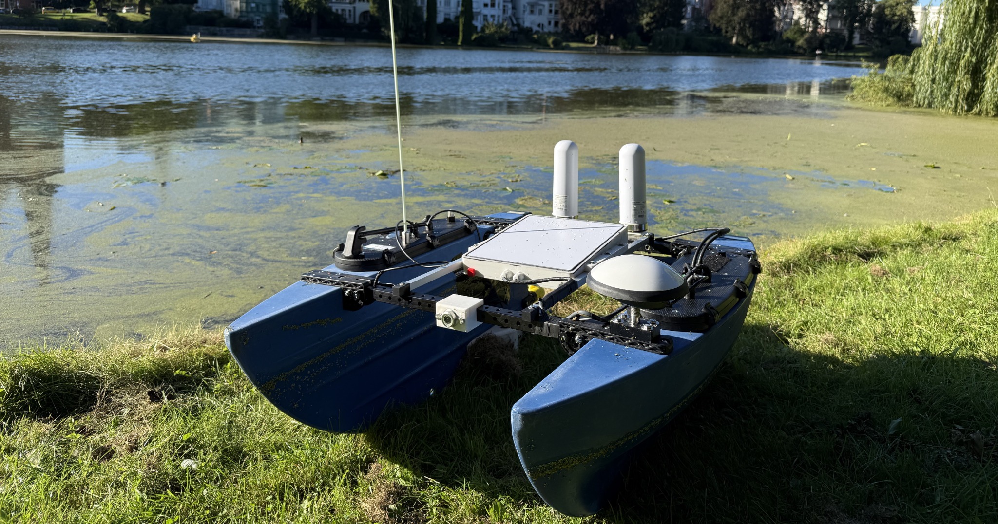 Das unbemannte Vermessungsboot NaviBlue am Ufer: blauer Katamaran-Rumpf mit GNSS-Antenne, zwei Kommunikationsantennen, Elektronikbox und Kamera