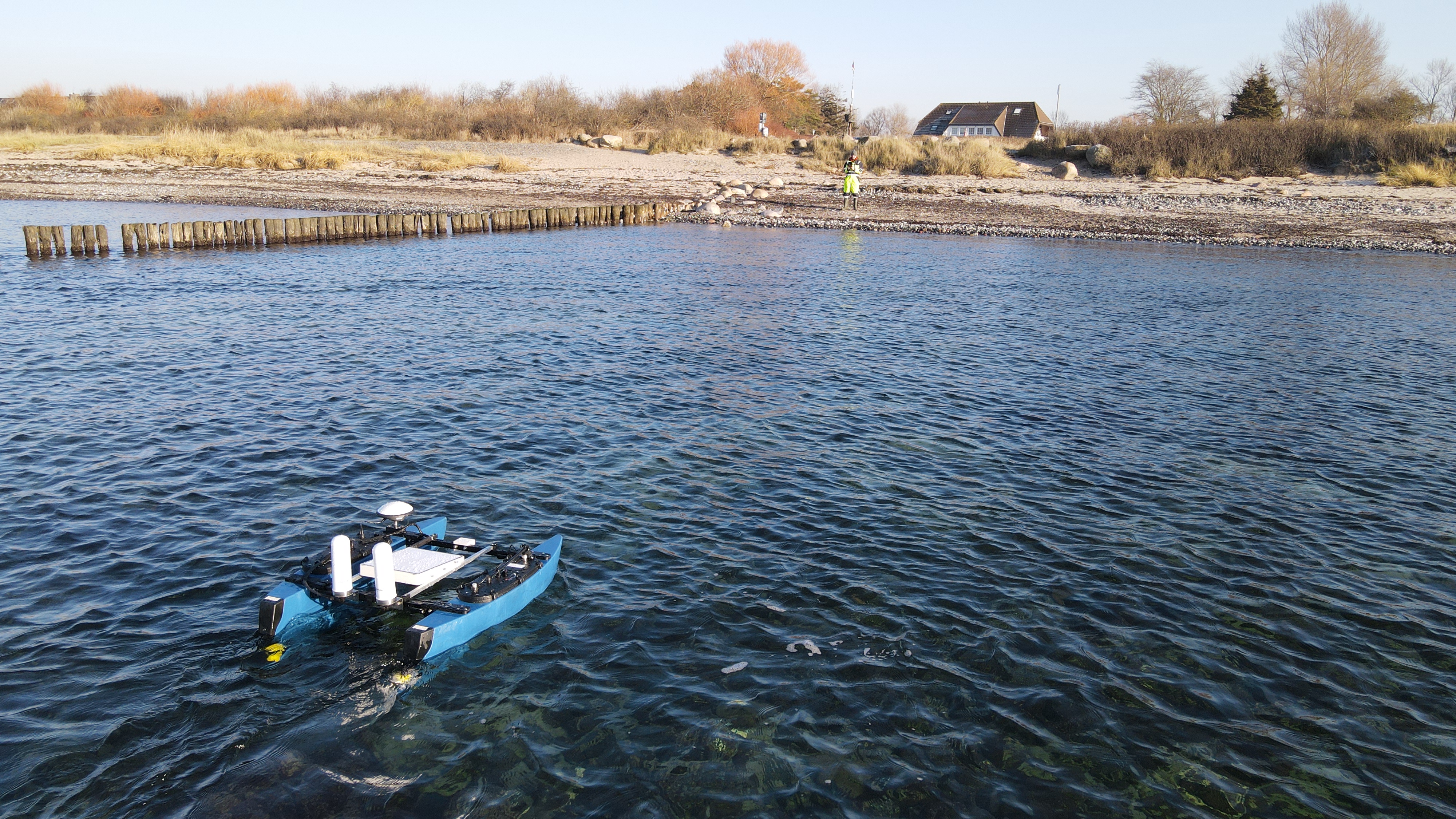 Das unbemannte Vermessungsboot NaviBlue im flachen Wasser vor einem Ostseestrand mit Buhnenreihe, am Ufer ein Vermesser in Warnkleidung